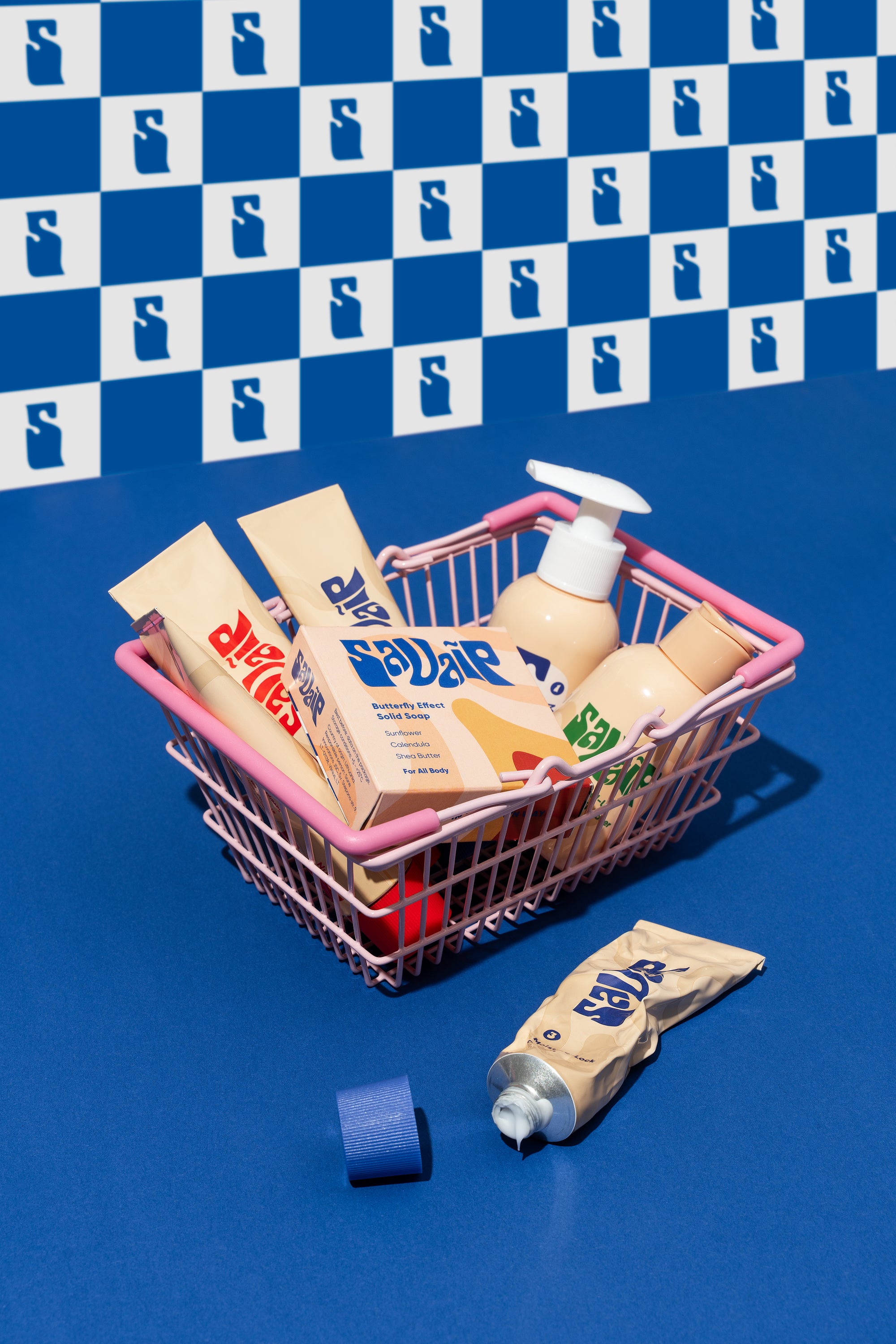 Miniature shopping basket filled with Savaip skincare and beauty products against a blue background with a repeated 'S'  Savaip Skin pattern; an open Savaip Moisture-Lock cream tube lies next to the basket.