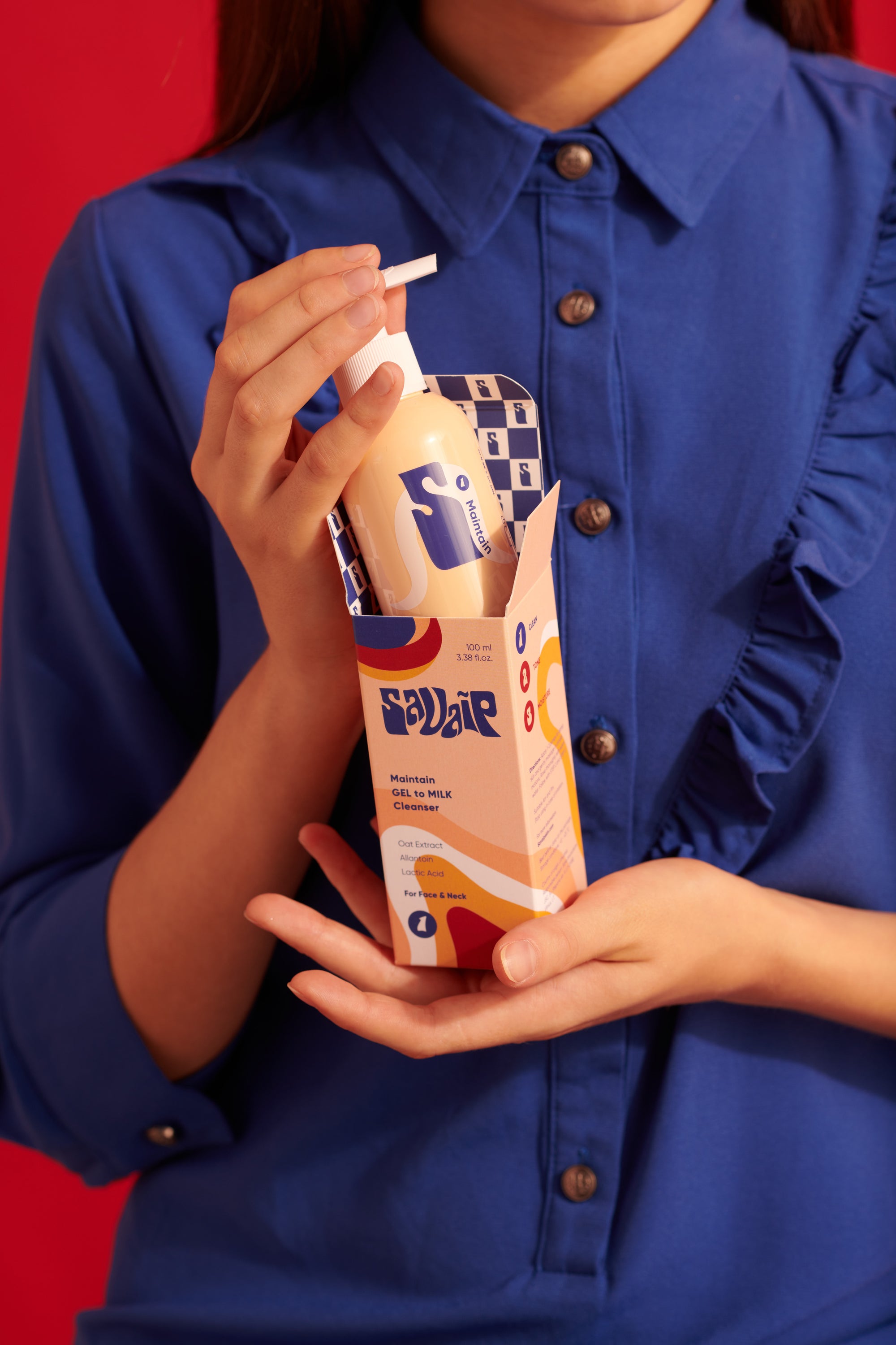 A person in a blue shirt holding a peach-colored Savaip skincare bottle labeled "GEL to MILK Cleanser" with details like Oat Extract, Allantoin and Lactic Acid on the packaging, set against a vibrant red background.