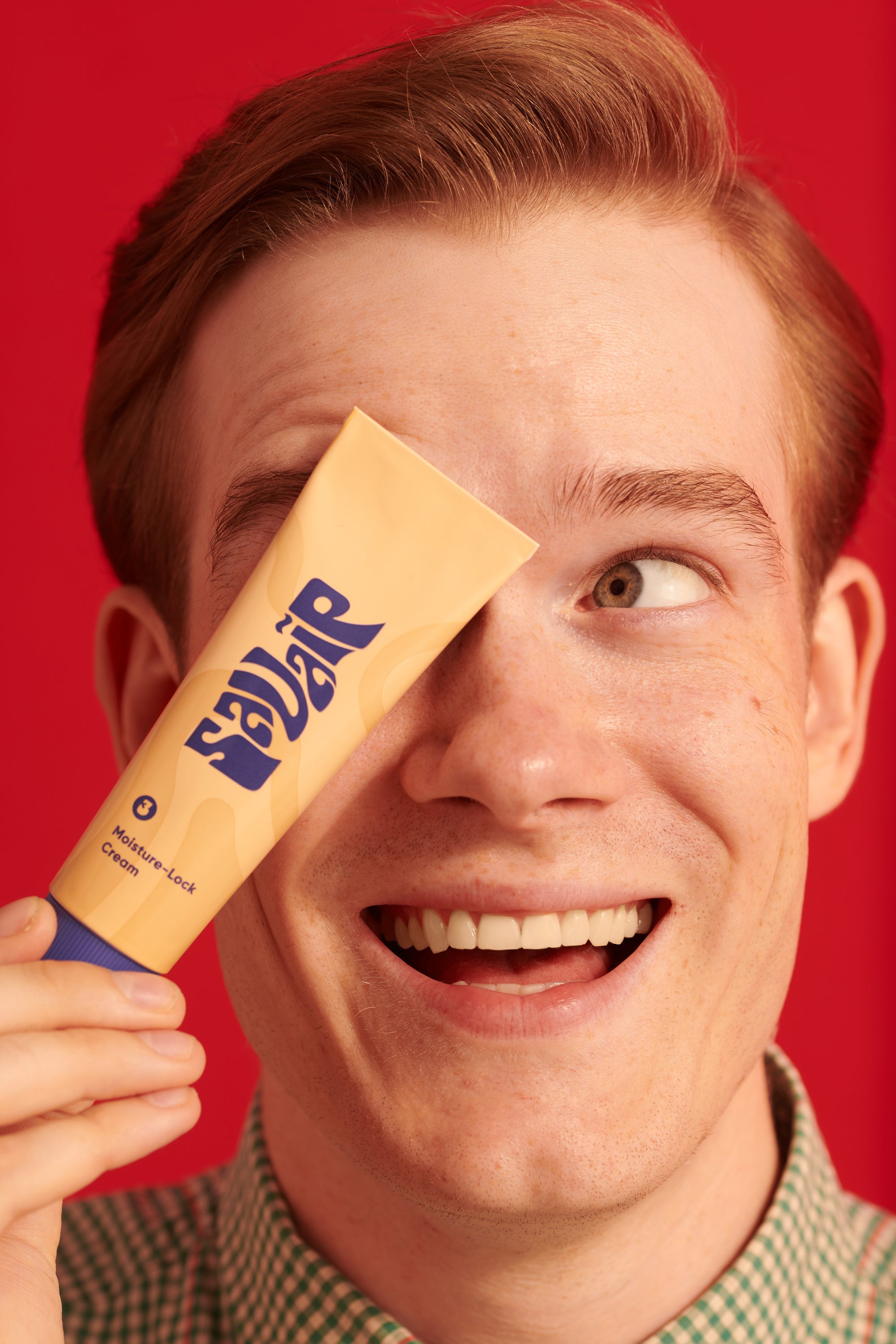 A young man with red hair smiling and holding up a yellow tube of Savaip Moisture-Lock Cream against a red background.