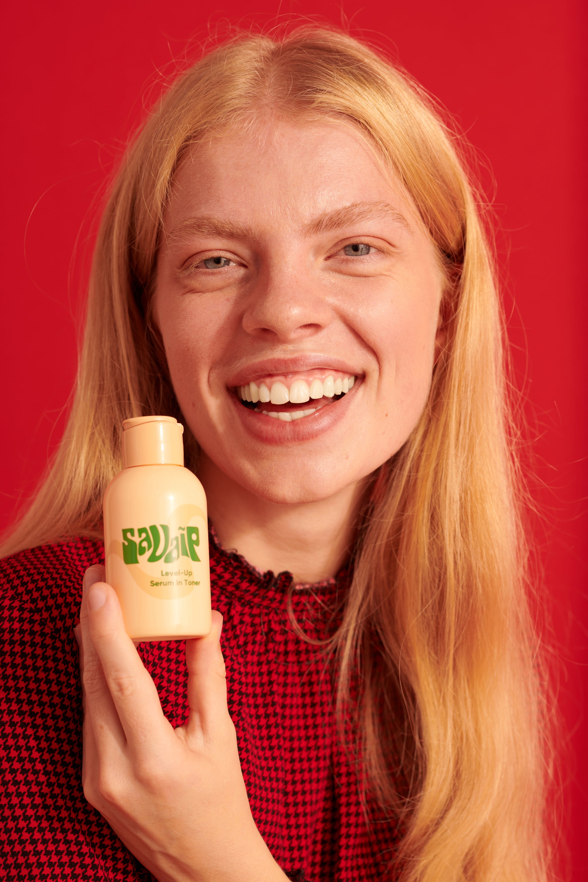 A person with long blonde hair and a joyful expression, holding a beige Savaip skincare bottle labeled Level-Up Serum in Toner, wearing a red houndstooth-patterned top against a red background.
